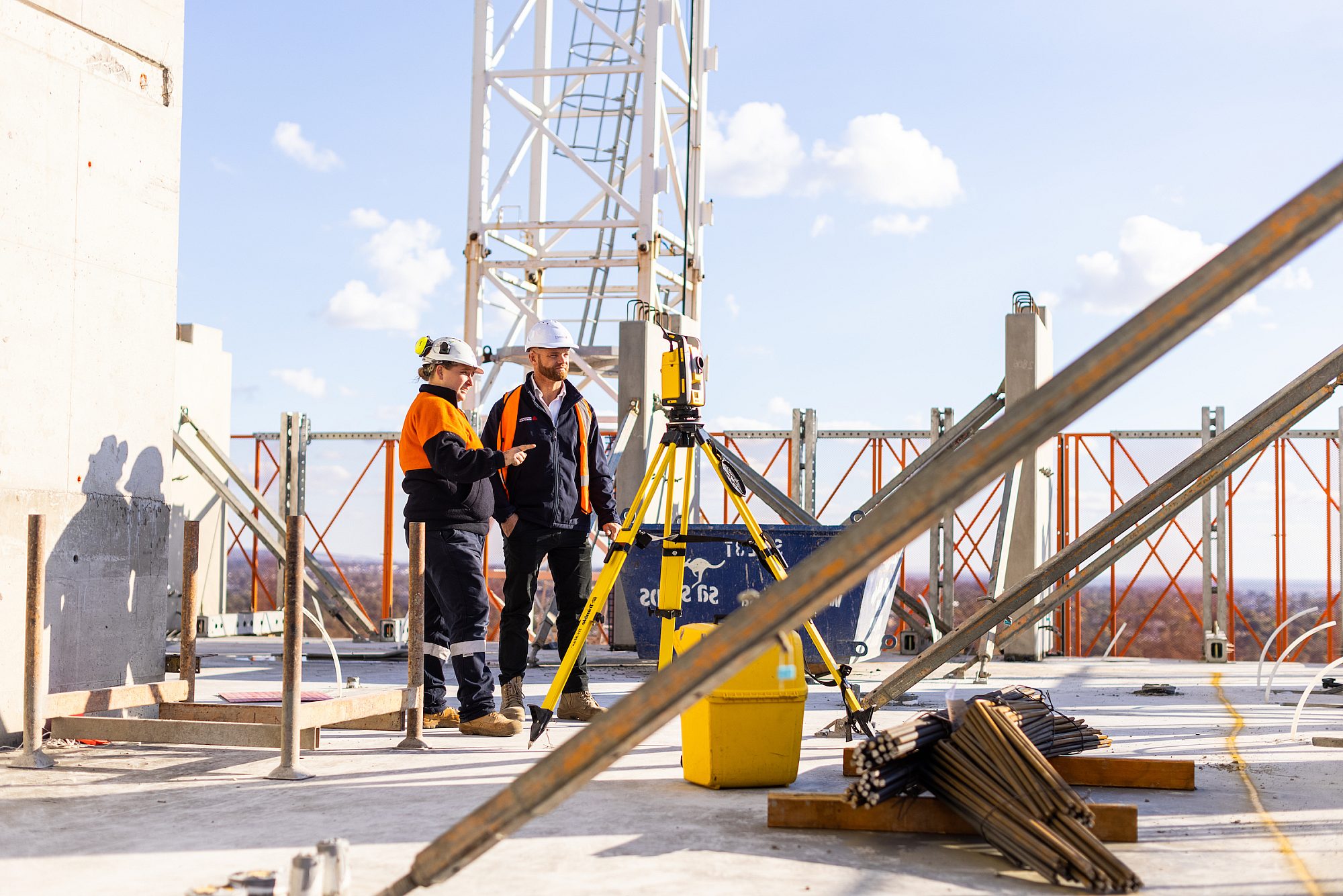 Two work people wearing hard hats and high vis clothing on a rooftop showing construction.